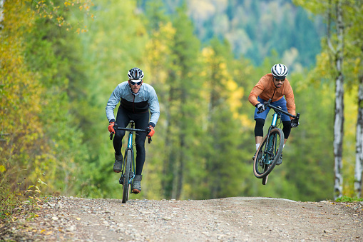A man catches some air while riding his gravel bicycle on a gravel, forestry road. Gravel bicycles are similar to road bikes but have oversized tires for riding on rough terrain.