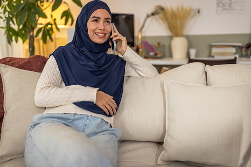 Photo of Arabian young woman is sitting on sofa at home and using her phone.