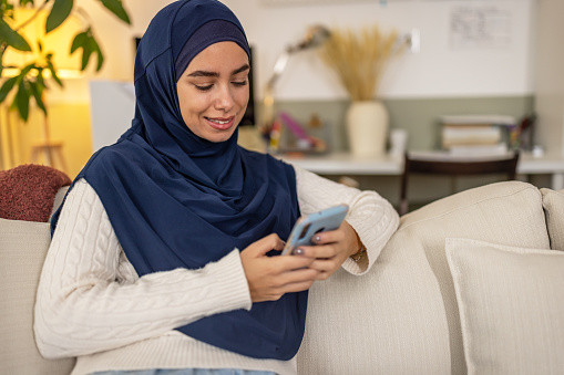 Portrait of young happy muslim woman in blue hijab sitting on a sofa using mobile phone.