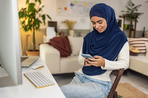 Portrait of young happy muslim woman in blue hijab using mobile phone at home.