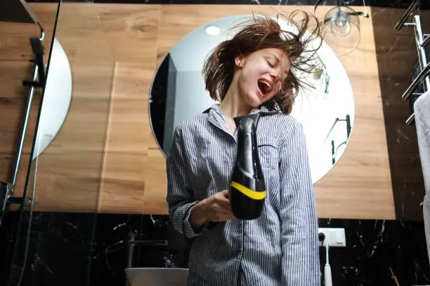 Concept of haircare and having fun in bathroom. Bottom view of cheerful beautiful woman singing while drying hair with blowdryer and using a hair dryer as a microphone at home stylish bath interior.