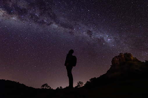 The silhouette of an adult man standing looking up at the sky while watching the milky way. He was on a night walk near Torre de Pedra, in a rural region of Ribeirão Claro, Paraná State, Brazil. The impression one has is that the stars come out of the Stone Tower, as if stars come out of a chimney.