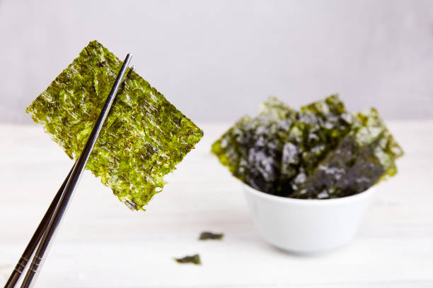 Chopsticks with a piece of crispy dried seaweed. Nori, Japanese edible seaweed in a plate. Chopsticks with a piece of crispy dried seaweed. Nori, Japanese edible seaweed in a plate. A healthy snack. nori edible seaweed sheet stock pictures, royalty-free photos & images