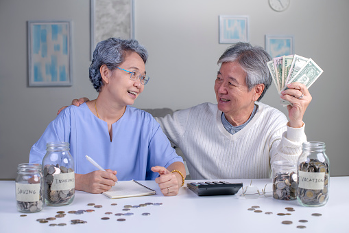 Savings concept. Asian old elderly senior couple grandparents husband and wife saving money, economizing pension, putting coin into money box, thinking over pension plan