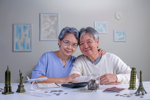 Portrait of happy Asian senior couple plans to save money for a trip around the world, looking at the camera.Travel budget concept, passport monument model and aircraft toy in the picture.