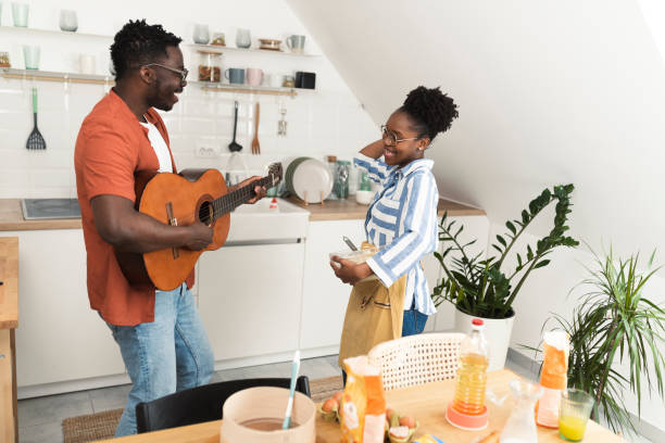 African man cheering up his wife and playing a guitar for her while she's making cookies stock photo