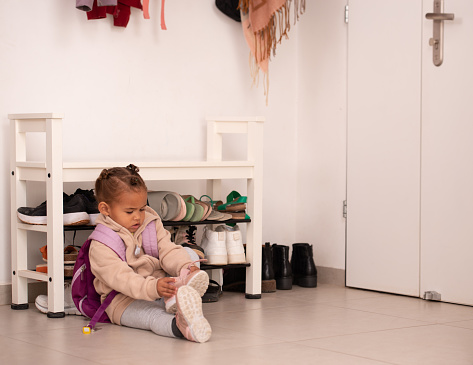 Determined little baby girl trying to put on her shoes inside at home entrance room before going to kindergarten. The child sitting on a tiled floor next to a shoe storage bench.