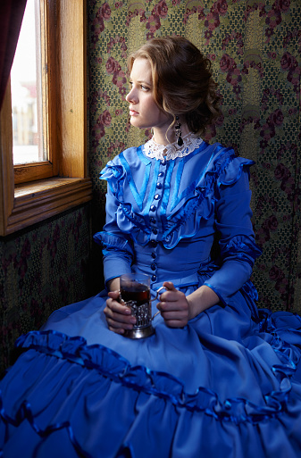 Young woman in blue vintage dress late 19th century sitting in coupe of retro railway train, looking out the window and drinking tea