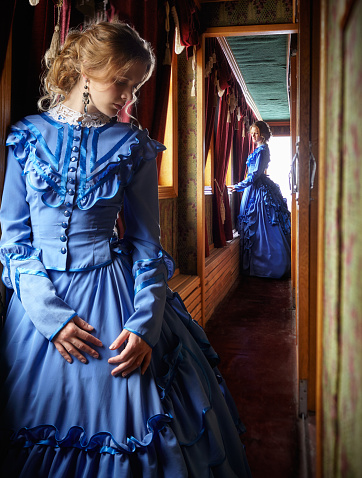 Young woman in blue vintage dress late 19th century standing near window in corridor of retro railway vehicle
