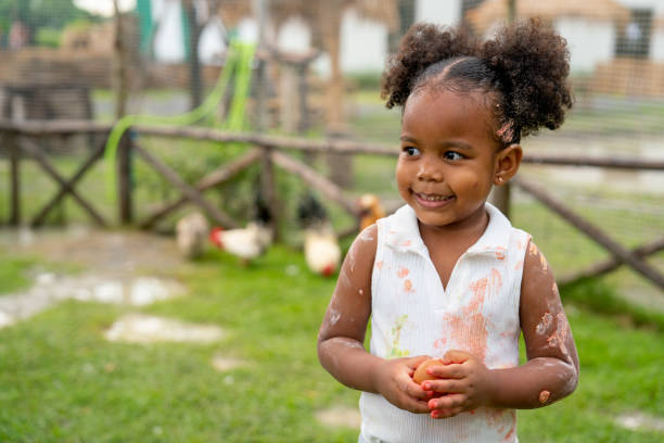 絵の具で遊んだ後、アフリカ系アメリカ人の小さな女の子は、慎重にサポートして卵を手に持ち、農場の左側を見ると、笑顔で幸せそうに見えます。 - multi ethnic group teenage girls little boys child ストックフォトと画像