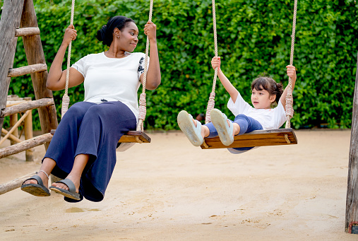 Little Caucasian girl talk to African American woman during play swing in playground of public park and they look enjoy and relax from outdoor activity.