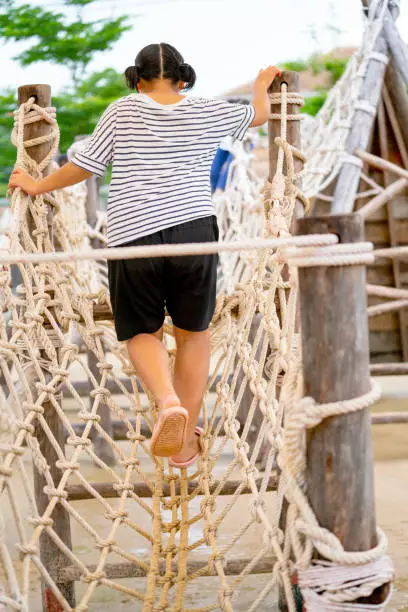 Back of Asian little girl walk along net bridge as plaything in playground of public park in concept of good activity for children to support development and creation of them.