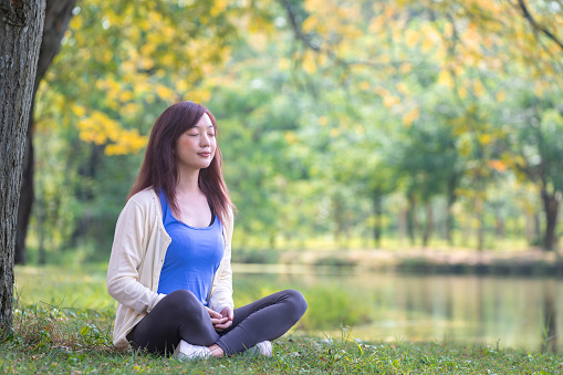 Woman relaxingly sitting and practicing meditation in the public park to attain happiness from inner peace wisdom under the tree in the summer