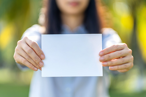Woman's hands holding a empty white paper standing against nature green background. Blank white card for design mockup