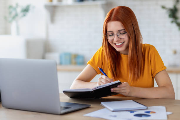 Happy young caucasian woman student using laptop computer for online education, watching the webinar, taking notes in notebook, studying, distance learning at home stock photo