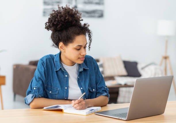istock Concentrated young African American woman student using laptop computer for online education, taking notes in notebook, studying, distance learning at home 1441117582