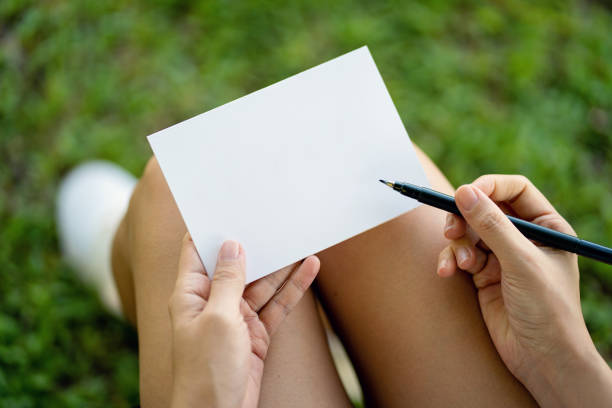 Woman's hand holding a pen and empty white paper sitting on grass field background. Blank white card for design mockup. High angle view.