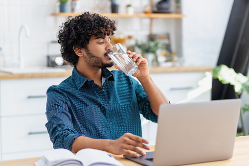 Young man freelancer or entrepreneur working at home office, taking a break, drinking refreshing water. Healthy lifestyle, recuperation concept