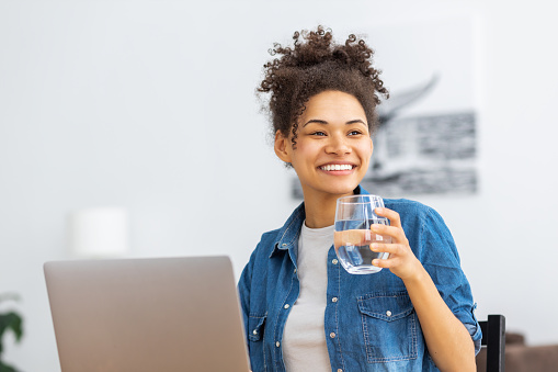 Healthy lifestyle, recuperation concept. Happy African American woman working in office using laptop holding glass of water smiling looking away