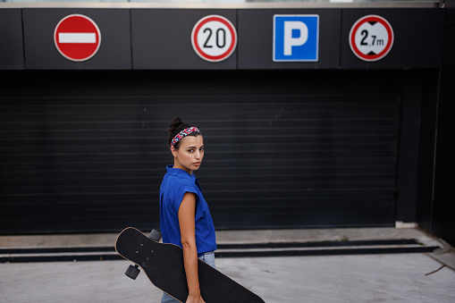 Beautiful young woman standing and holding longboard in hand while looking at camera in the city