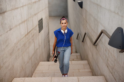 Young skate woman carrying longboard and walking up the stairs in the city