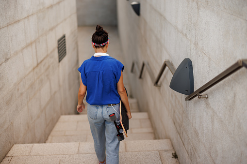 Young skate woman carrying longboard and walking down the stairs in the city, back view