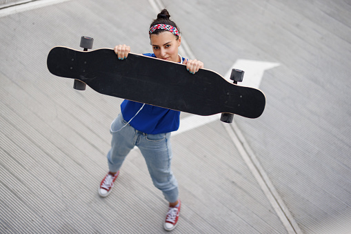 High angle view of young skate woman holding longboard and looking at camera in the city