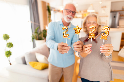 Senior couple having fun spending winter holiday season together at home, holding small golden balloons shaped as numbers 2023, representing upcoming New Year