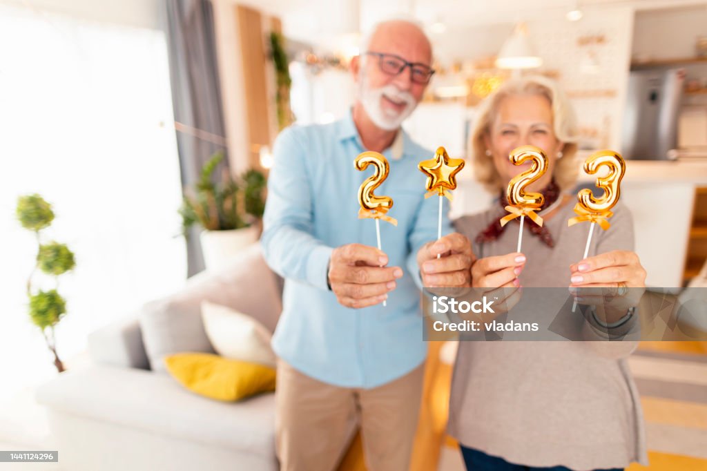 Senior couple holding numbers 2023 for New Year home party Senior couple having fun spending winter holiday season together at home, holding small golden balloons shaped as numbers 2023, representing upcoming New Year Senior Adult Stock Photo