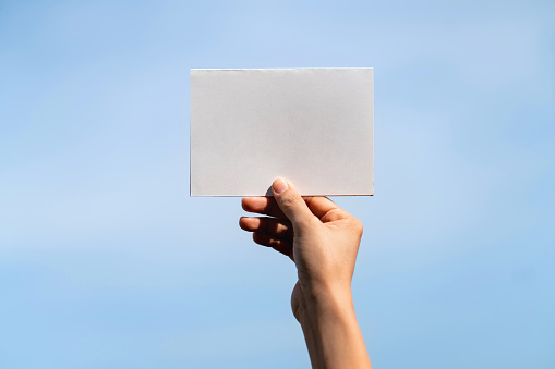 Woman's hand holding a empty white paper against blue sky background. Blank white card for design mockup.