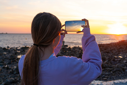 Girl tourist woman taking a smartphone photo of the sunset in the sea rear view with ponytail blond hair