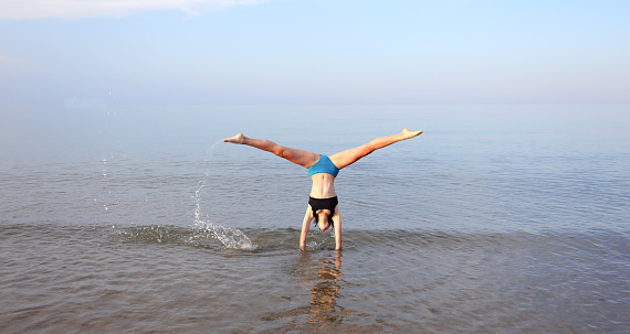 young girl performs gymnastic exercises with her head upside down on the seashore in summer