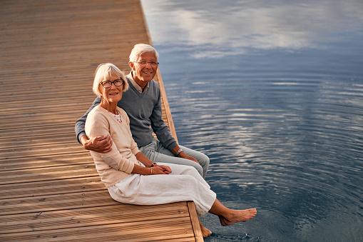 An elderly, intelligent, gray-haired couple in love are sitting on a pier in a seaport, having a romantic time admiring the seascape.