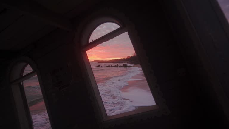 View from flooded leaning wooden house room window washed by sea waves at coastline on spectacular pink sunset. Vintage rickety building sinking in water on sandy beach on dreamy sunrise skyline.