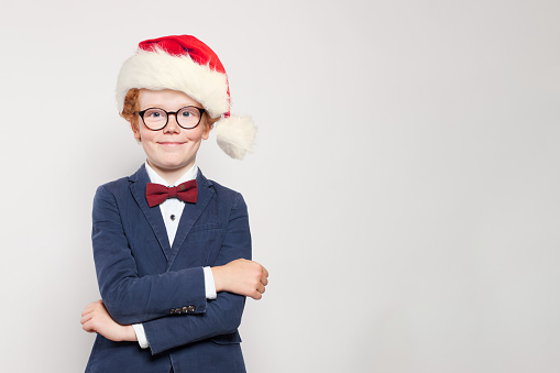 Happy red haired child boy in Santa hat standing against white wall background