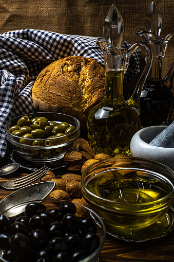 fresh food on the table. the photo represents oil, vinegar, salt, green olives, black olives, carrots, celery, bread and potatoes. the background is made from jute and a red and white checkered tablecloth that evokes tradition. the food is stored in glass bowls, wicker baskets and cruets. dark mood