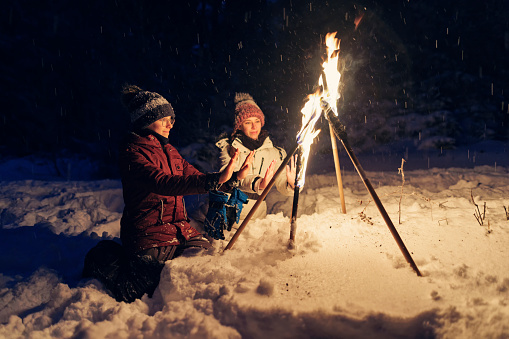 Kids hiking in beautiful winter forest at night. Kids are warming hands with flaming torches stuck in snow.\nShow with Canon R5