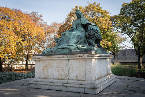 The impressive sculpture symbolizes the unification of Geneva with Switzerland in 1814.\nTwo women in bronze attire – the Republic of Geneva and Helvetias – holding each other entwined at the waist. Perched on their pedestal, they look north towards Switzerland.\n\nThe National Monument is located in the Jardin Anglais, facing the lake. It was inaugurated in 1869. With this work, the sculptor Robert Dorer symbolised Geneva’s integration into the Swiss Confederation on 12 September 1814 with two women each carrying a sword and shield.