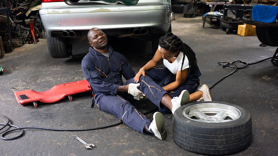 African American woman mechanic helped a black engineer man with knee pain due to work lie on the garage floor.