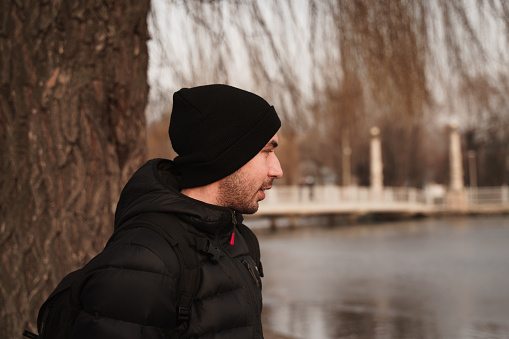 A side view of a young Ukrainian bearded male wearing a black beanie and a winter jacket in a park