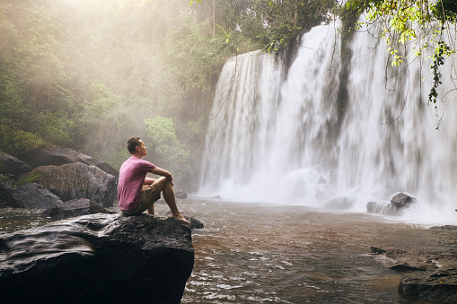 Man sitting on rock in front of high waterfall in mountains in tropical landscape in Camobodia.