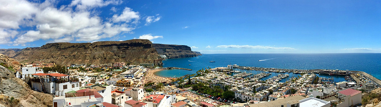Aerial view, in panoramic format, of Puerto de Mogan, in Gran Canaria, Spain, showing, buildings, beach, bay, sea and nearby hills on the coast.