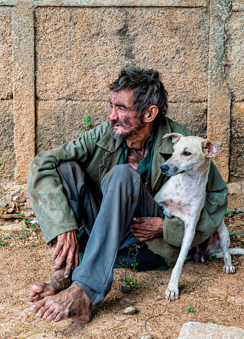 A vertical shot of a black homeless man sitting outdoors with a dog