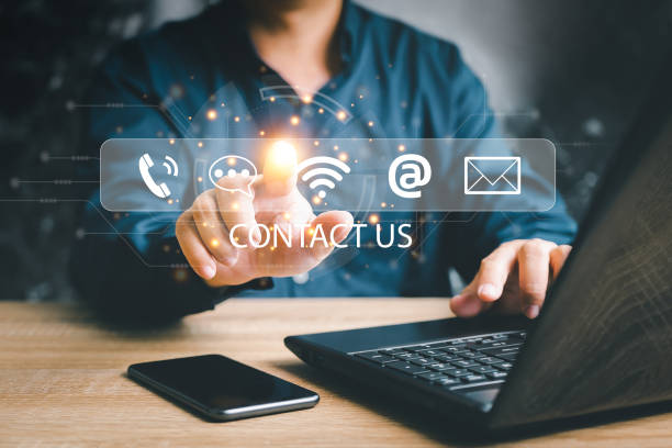 istock Contact us or the customer support hotline people connect. Businessman point virtual screen on contact icons and using laptop smartphone on wood table 1441181069