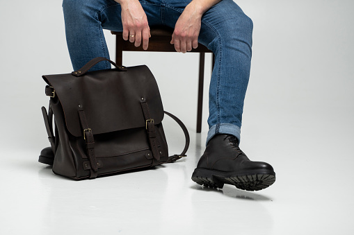 Man in a blue jeans and black boots sits on a chair with a brown men's shoulder leather bag for a documents and laptop on a white floor. Mens leather satchel, messenger bags, handmade briefcase