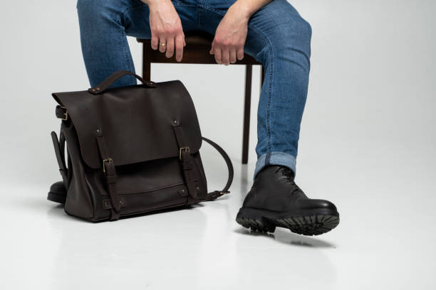 Man in a blue jeans and black boots sits on a chair with a brown men's shoulder leather bag for a documents and laptop on a white floor. Mens leather satchel, messenger bags, handmade briefcase. stock photo