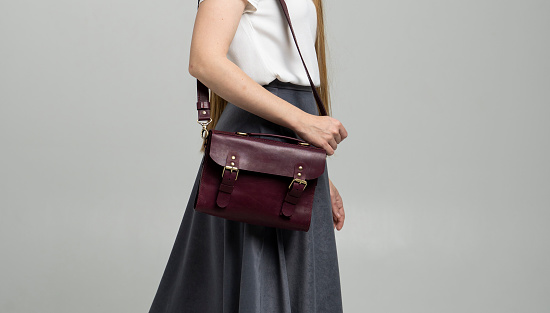 Small red leather bag over a woman shoulder on a white background. Shoulder handbag. Woman in a white shirt and grey skirt and with a ted handbag. Style, retro, fashion, vintage and elegance