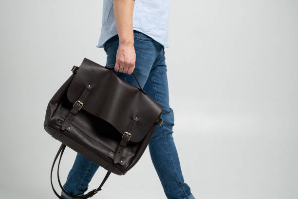 Brown men's shoulder leather bag for a documents and laptop holds by man in a blue shirt and jeans with a white background. Satchel, mens leather handmade briefcase. stock photo