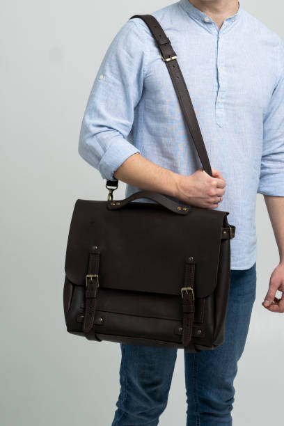Brown men's shoulder leather bag for a documents and laptop on the shoulders of a man in a blue shirt and jeans with a white background. Satchel, mens leather handmade briefcase. stock photo