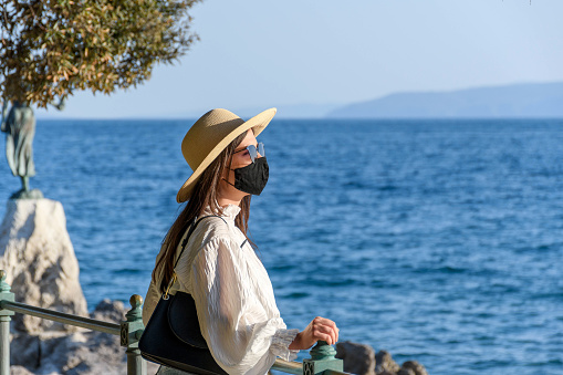 Side view of female tourist with black protective mask and sunglasses standing on shore of sea during covid epidemic in Opatija, Croatia.